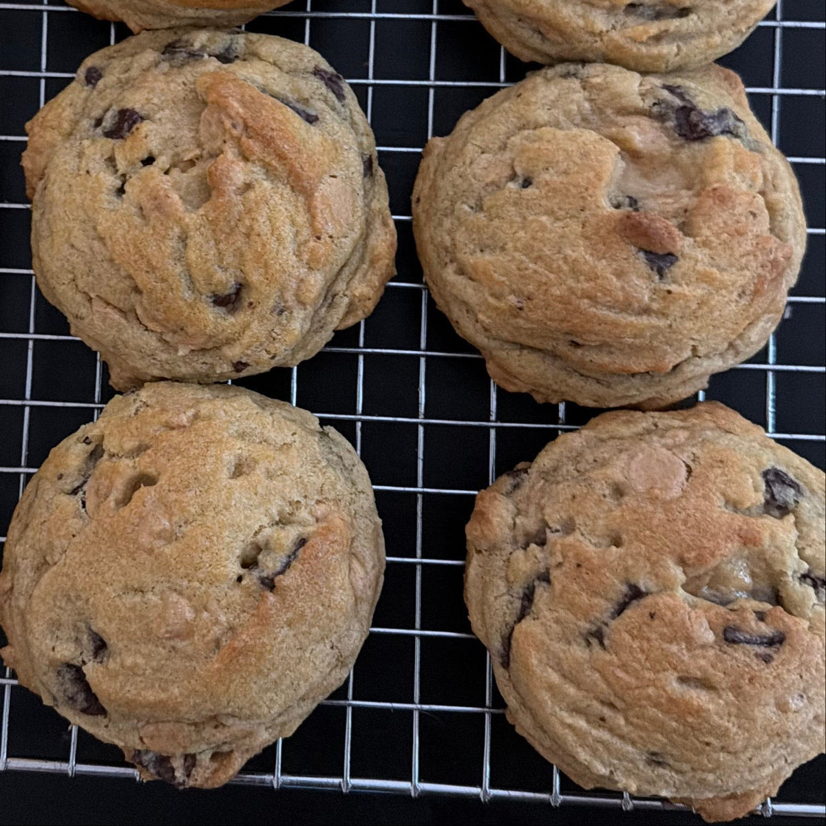 Nine chocolate chip cookies cooling on a wire rack over a black surface.